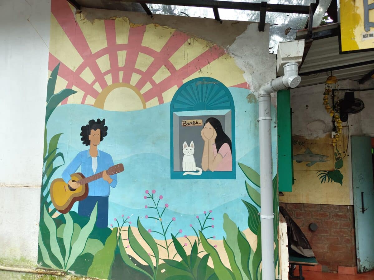 The image shows a colorful mural with a person playing a guitar, a framed picture featuring another person and a cat, surrounded by plant motifs.