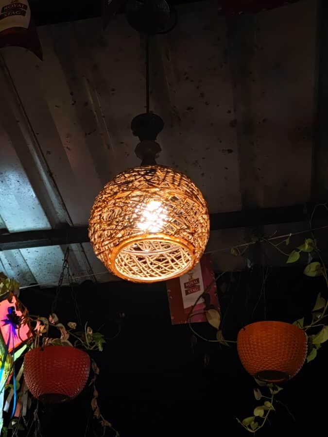 An illuminated wicker pendant lamp hangs from a ceiling, with surrounding decor of greenery and other colorful, textured lamps against a dark background.