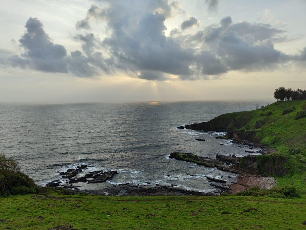 This image shows a serene coastal landscape with green grassy cliffs, layered rock formations at the shoreline, and a sunbeam piercing through the clouds over the sea.